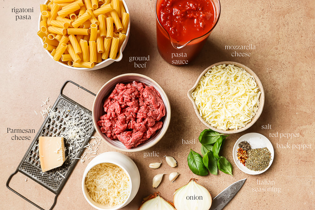 Top-down view of ingredients for Baked Rigatoni with Ground Beef, including uncooked rigatoni, ground beef, pasta sauce, shredded mozzarella, Parmesan with a grater, garlic cloves, basil leaves, halved onion, and assorted seasonings.