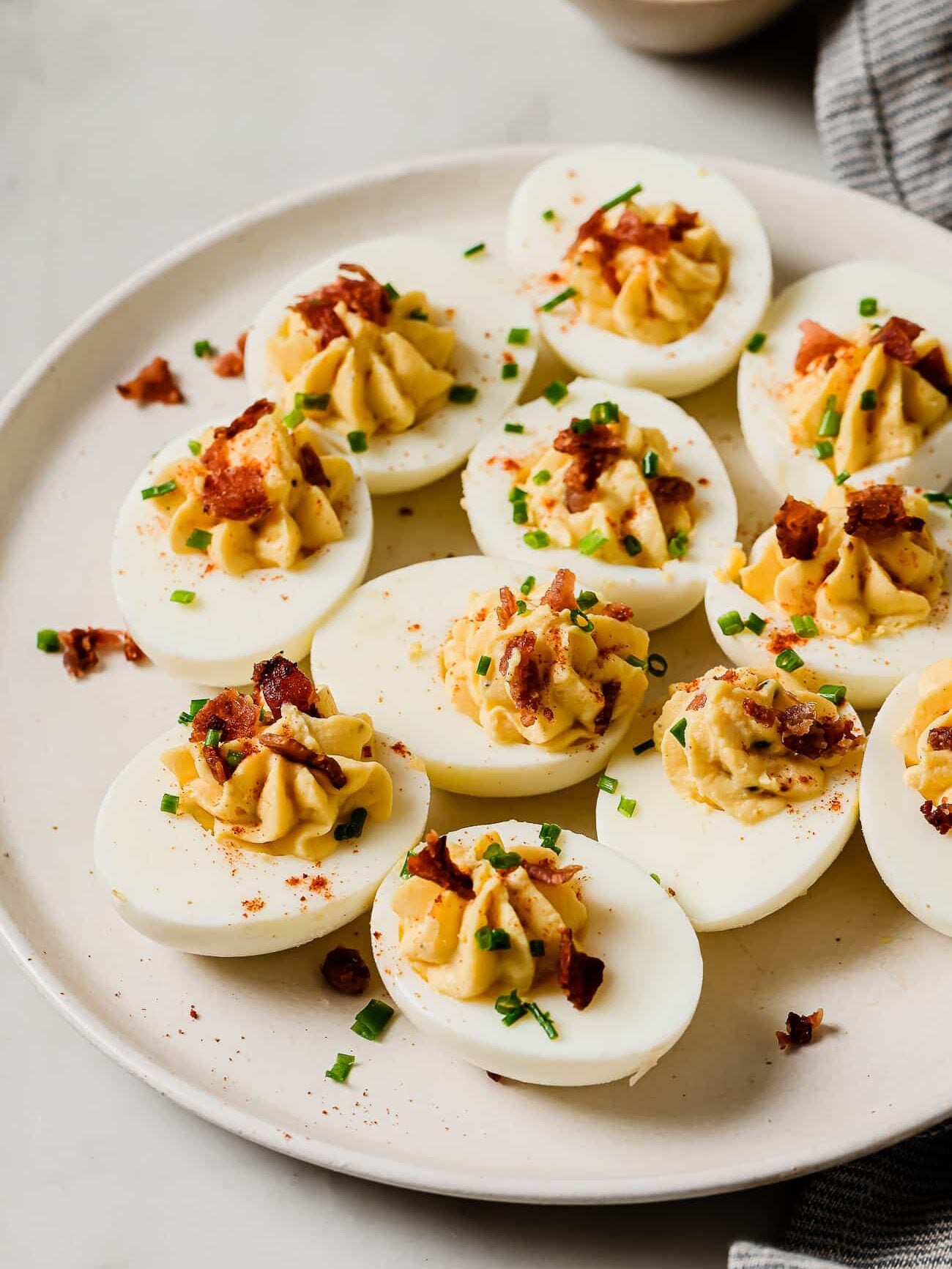 A plate of Easy Deviled Eggs, topped with crumbled bacon and chopped chives, sits on a white surface. A bowl of herbs and a striped napkin are partially visible in the background.