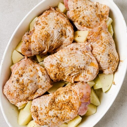 Raw seasoned chicken thighs arranged on top of chopped potatoes in a white oval baking dish, ready to be cooked.