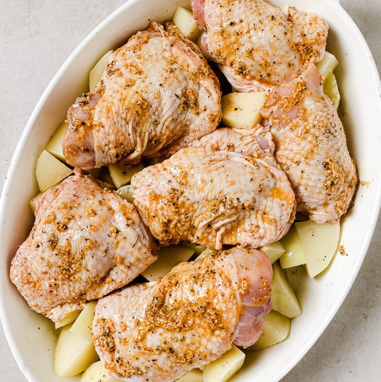 An overhead view of marinated chicken thighs over diced potatoes in a white baking dish.