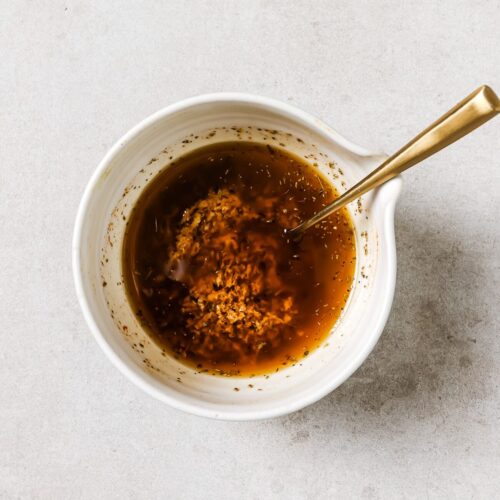 A white bowl filled with a brown liquid marinade, containing visible minced garlic and herbs, with a gold spoon resting inside, set on a light-colored surface.