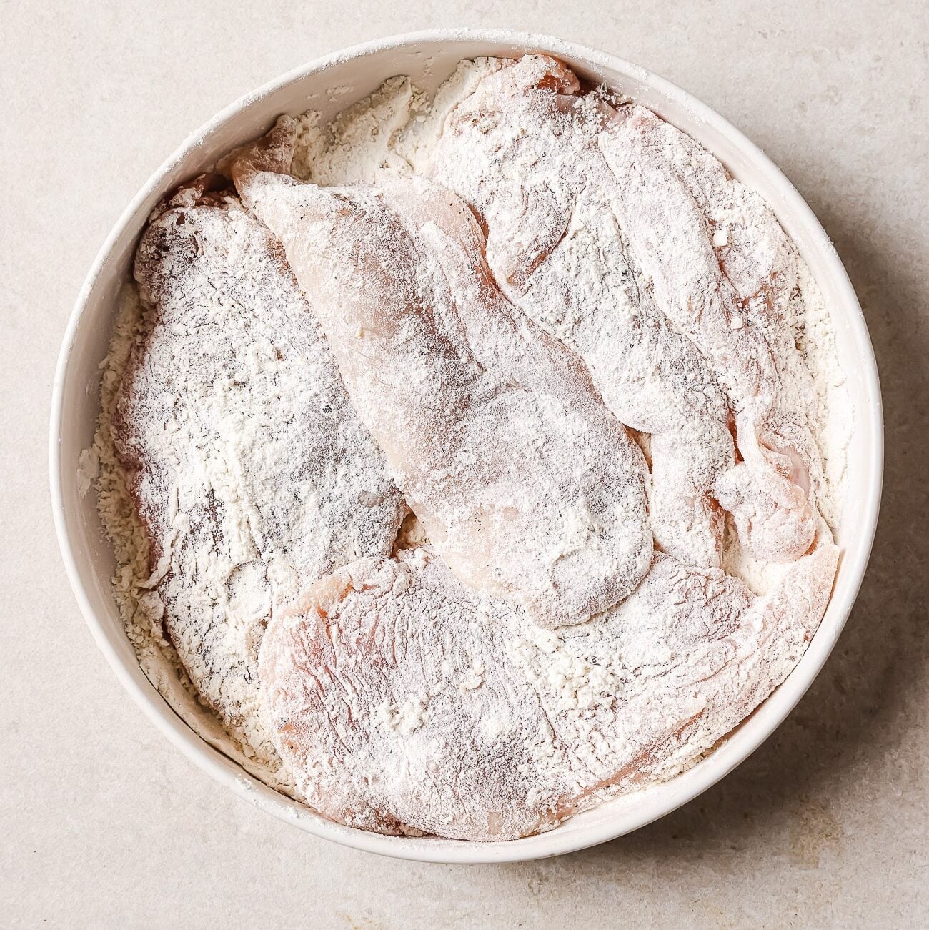 A large white bowl full of chicken cutlets dredged in flour.