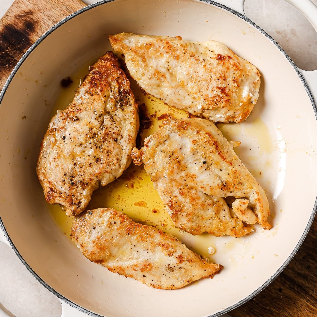 An overhead view of four seared chicken cutlets in a white skillet.