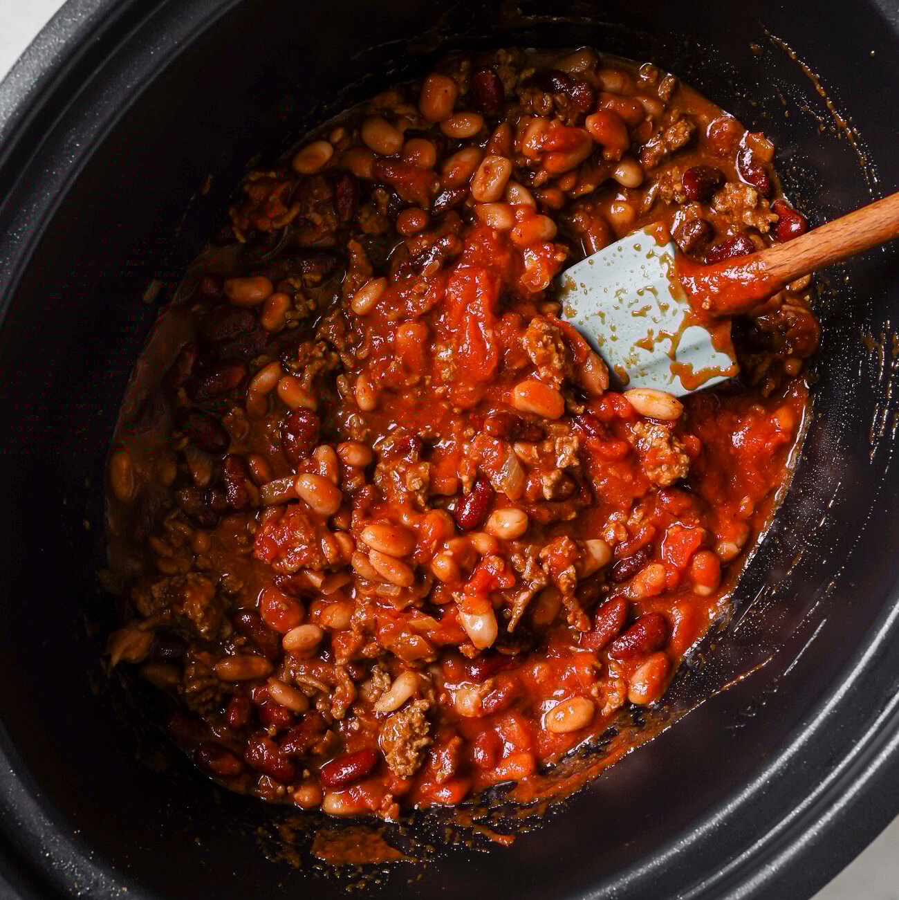 A slow cooker filled with a hearty Slow Cooker Chili Recipe—made of ground beef, kidney beans, white beans, and tomato sauce—being stirred with a blue spatula.