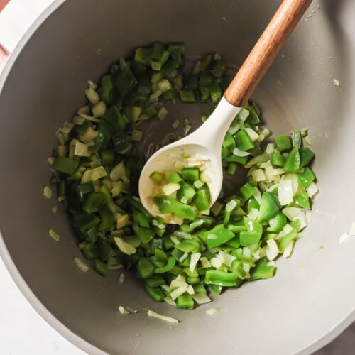 Chopped green bell peppers and onions being sautéed in a large pot with a white spoon, creating a flavorful base for White Chicken Chili.