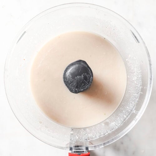 A food processor with a red handle contains a smooth, light beige White Chicken Chili mixture, viewed from above on a white marble surface.