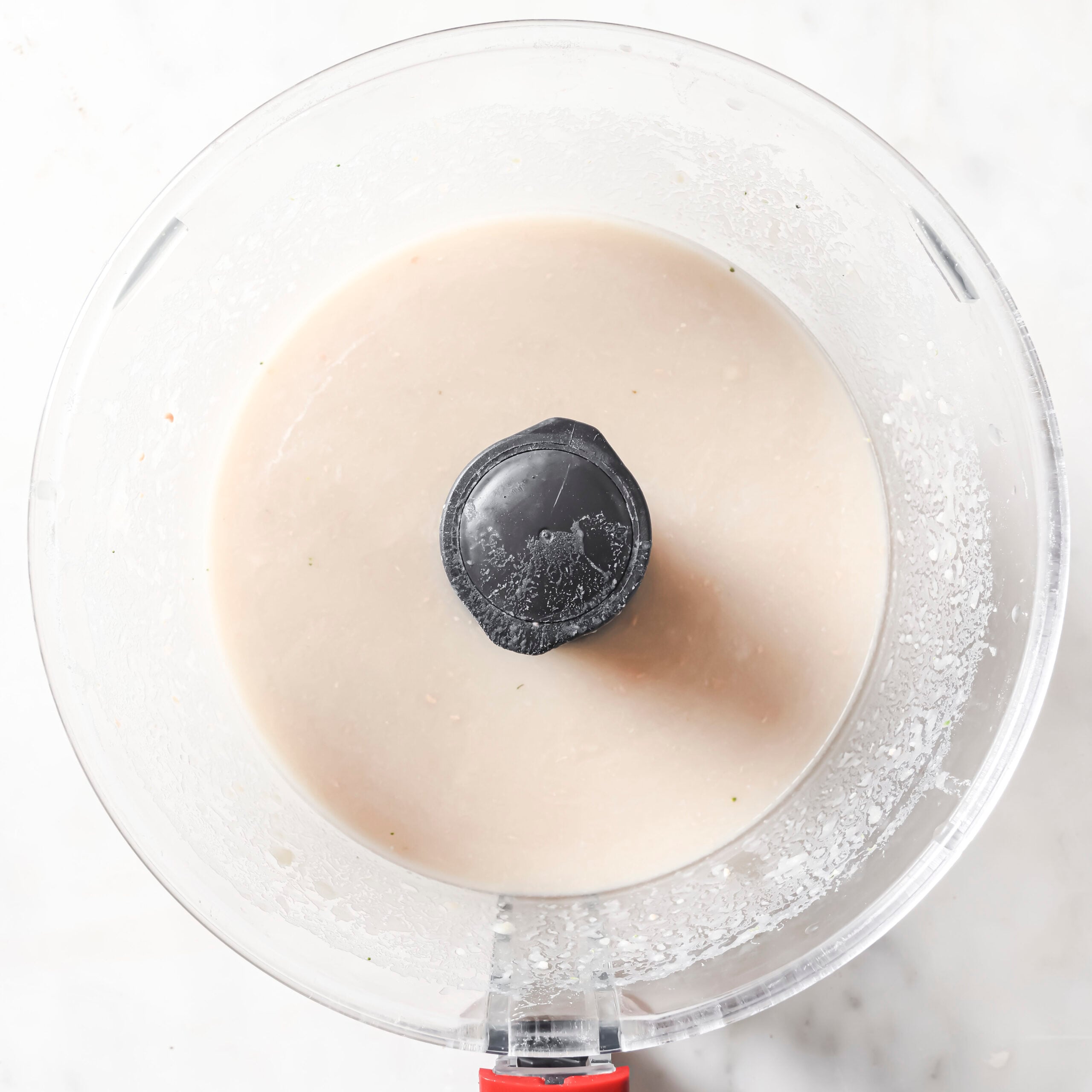 A food processor with a red handle contains a smooth, light beige White Chicken Chili mixture, viewed from above on a white marble surface.