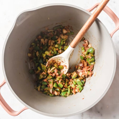 A pot on a marble surface contains chopped green bell peppers, onions, and spices being stirred with a wooden spoon—the perfect base for a hearty White Chicken Chili.
