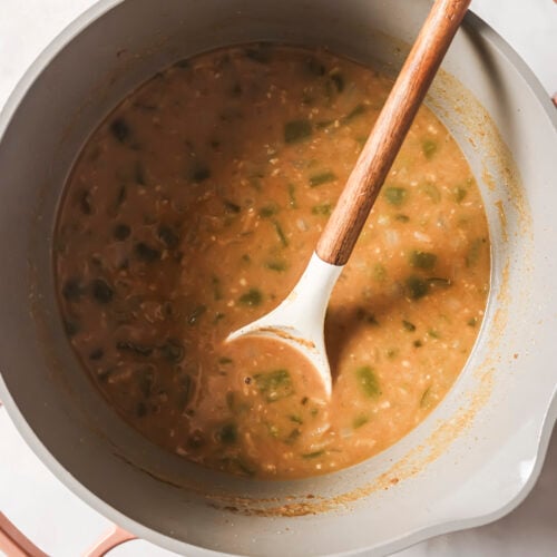 A pot filled with a thick, brown and green White Chicken Chili sits on a white surface. A wooden spoon with a white tip rests inside the pot, partially submerged in the hearty mixture.