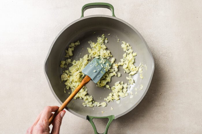 A hand stirs chopped onions in a large, light-colored pan with a green handle, using a spatula. The onions are being sautéed on a light-colored surface.