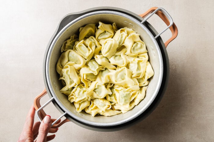 A hand holds a pot filled with uncooked tortellini pasta on a light-colored surface.