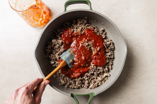 A hand stirs browned ground beef and chopped onions in a skillet with tomato sauce being mixed in. A glass with leftover sauce sits nearby on a light surface.