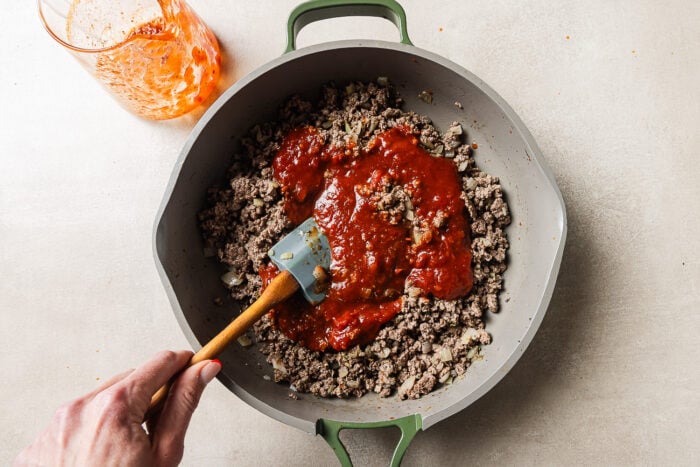 A hand stirs browned ground beef and chopped onions in a skillet with tomato sauce being mixed in. A glass with leftover sauce sits nearby on a light surface.