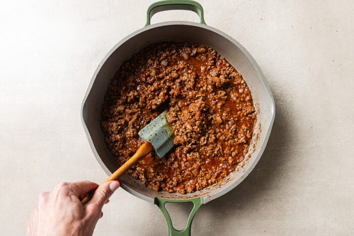A hand stirs seasoned ground beef and tomato sauce with a green spatula in a large gray pot on a light countertop.