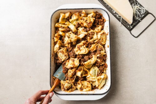 A hand holds a spatula in a baking dish filled with tortellini pasta and meat sauce. A block of Parmesan cheese and a grater sit on the surface nearby.