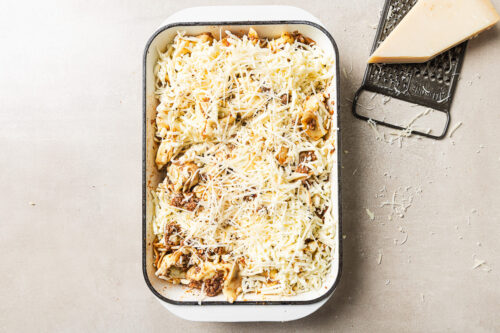 A rectangular baking dish filled with uncooked pasta topped with shredded cheese sits on a light surface. Next to it are a metal grater and a wedge of cheese, with cheese shavings scattered around.