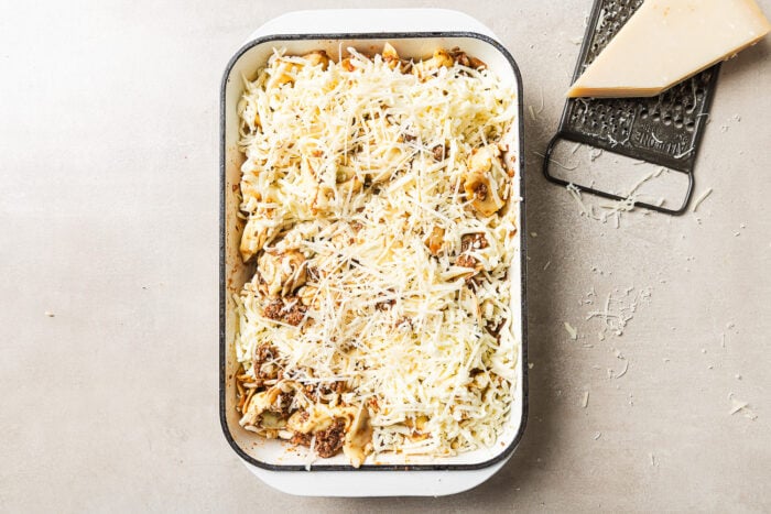 A rectangular baking dish filled with uncooked pasta topped with shredded cheese sits on a light surface. Next to it are a metal grater and a wedge of cheese, with cheese shavings scattered around.