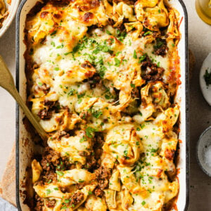 A baked pasta casserole in a white dish filled with cheese, ground meat, and herbs, topped with melted cheese and chopped parsley. A serving spoon is placed in the dish, ready for serving.