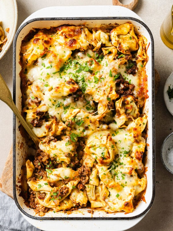 A baked pasta casserole in a white dish filled with cheese, ground meat, and herbs, topped with melted cheese and chopped parsley. A serving spoon is placed in the dish, ready for serving.