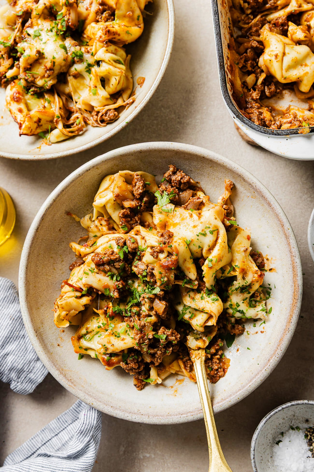 A bowl of tortellini pasta topped with a hearty meat sauce and fresh herbs, with a fork in the bowl. There are more servings of pasta in dishes nearby on a light-colored surface.