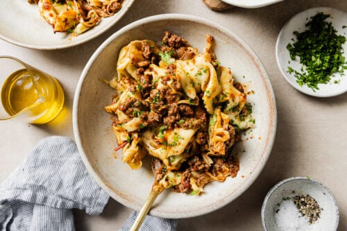 A bowl of pasta with crumbled ground meat, garnished with chopped herbs. Surrounding the bowl are a dish of salt and pepper, a small bowl of herbs, a glass container of olive oil, and a striped napkin.