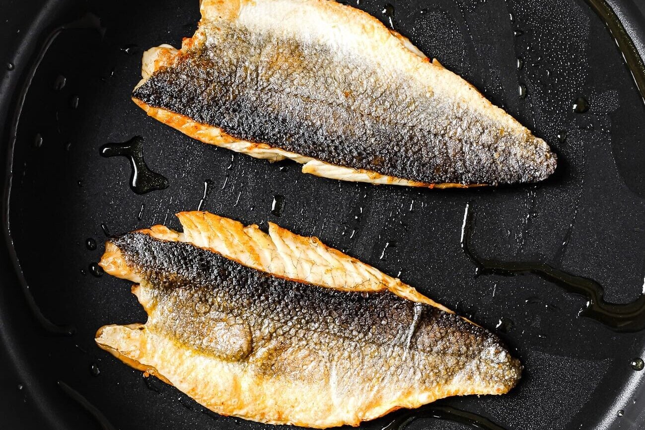 Two fillets of fish with crispy skin are cooking in a black nonstick pan with a drizzle of oil. The fish is golden brown and slightly curled at the edges.