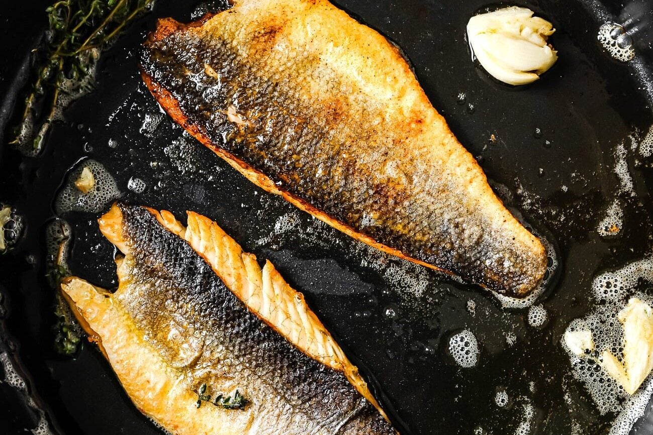 Two fillets of fish with crispy skin are being pan-fried in a skillet with bubbling butter, fresh herbs, and garlic cloves.