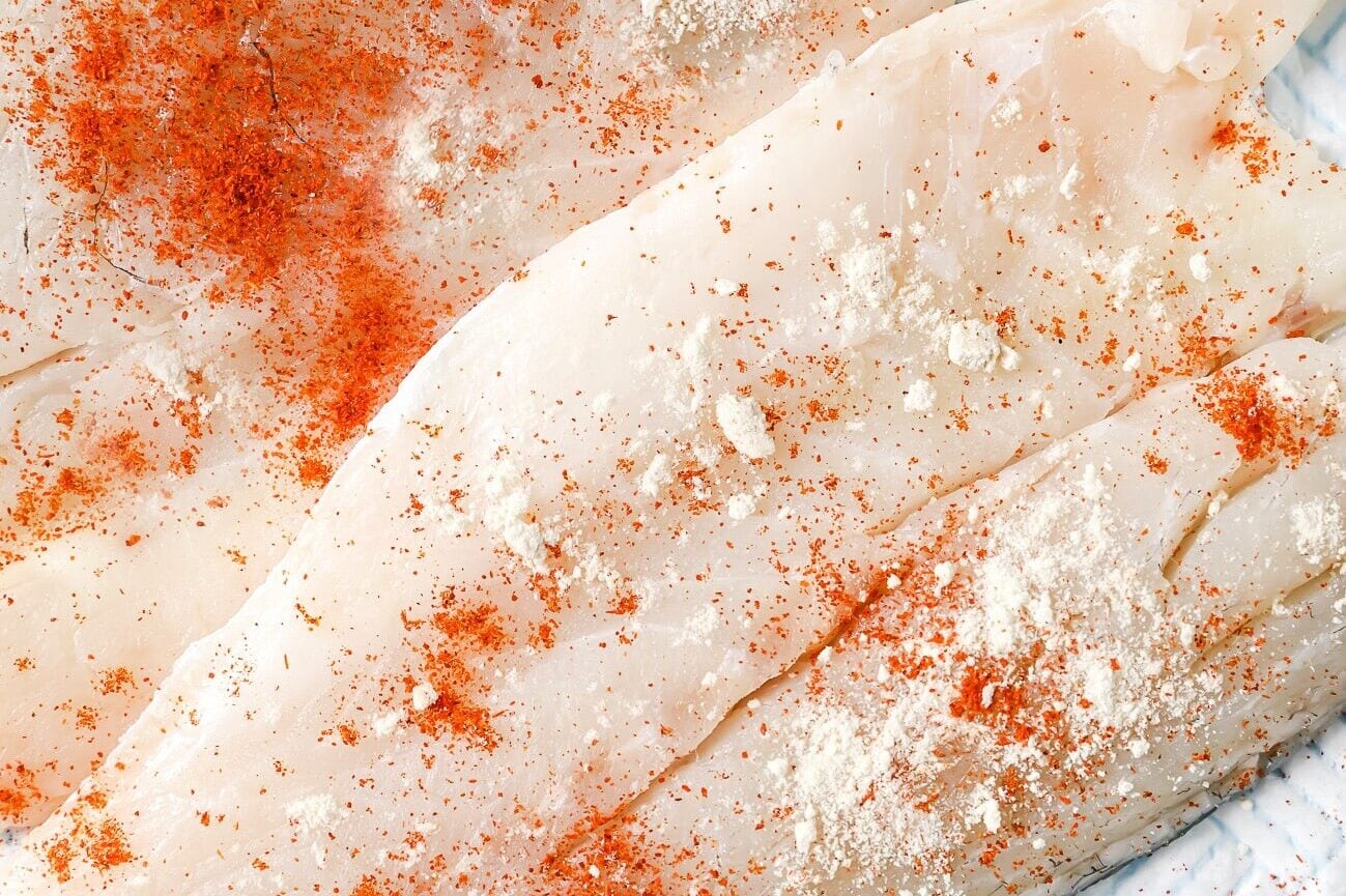 Raw fish fillets are sprinkled with red paprika and white seasoning, lying on a white textured surface, ready to be cooked.
