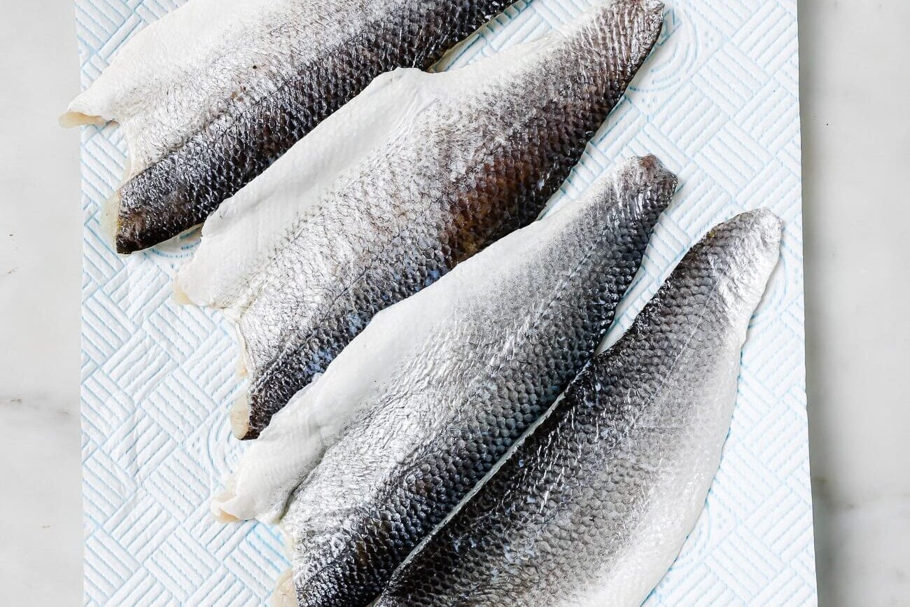 Four raw, skin-on fish fillets are arranged side by side on a textured white paper towel, set on a light marble surface. The fish skin appears silvery and shiny.