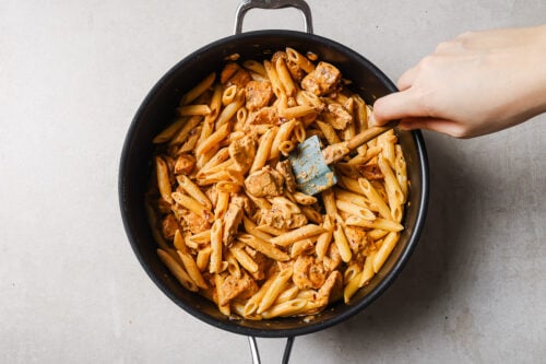 A hand stirs cooked penne pasta and chunks of chicken in a large black skillet with a blue spatula, set on a light-colored surface.