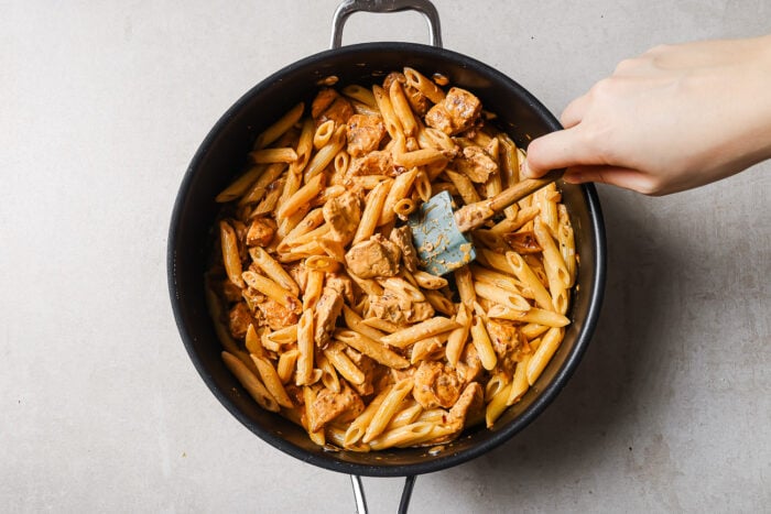 A hand stirs cooked penne pasta and chunks of chicken in a large black skillet with a blue spatula, set on a light-colored surface.
