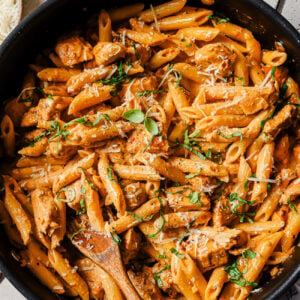 A large skillet filled with creamy penne pasta, pieces of chicken, fresh herbs, and grated cheese, with a wooden spoon for serving. Bowls of chopped herbs and grated cheese are nearby on the table.