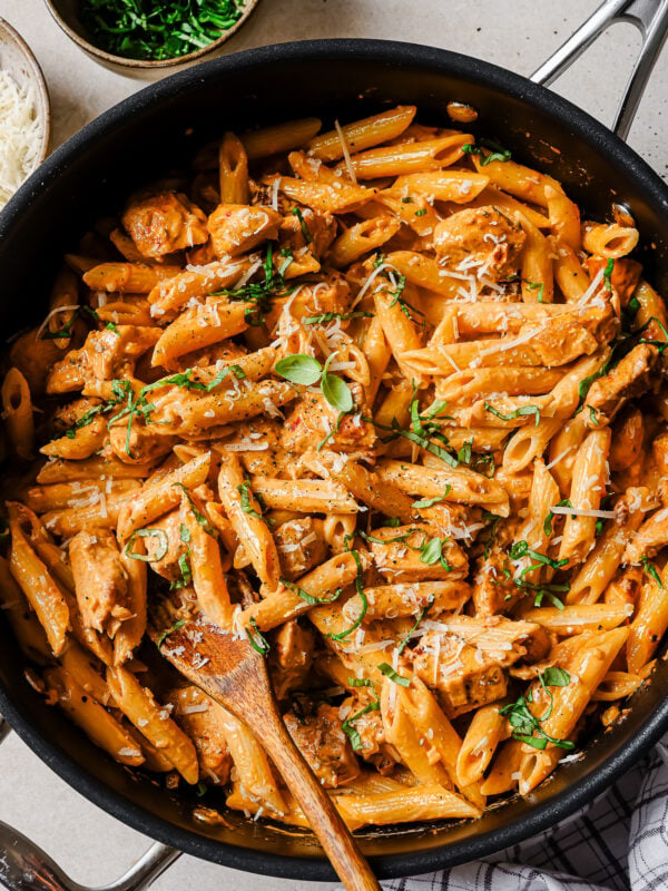 A large skillet filled with creamy penne pasta, pieces of chicken, fresh herbs, and grated cheese, with a wooden spoon for serving. Bowls of chopped herbs and grated cheese are nearby on the table.