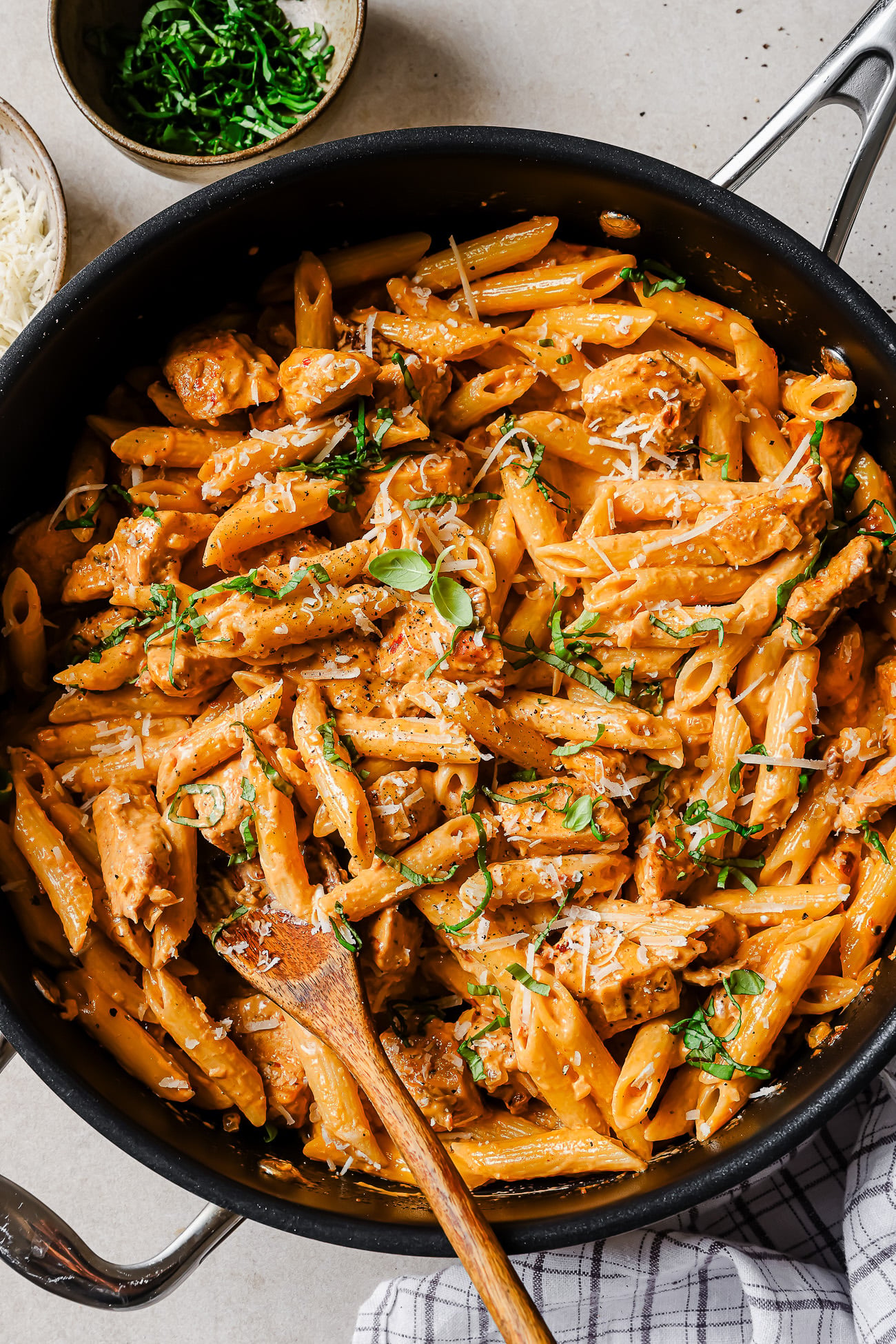 A large skillet filled with creamy penne pasta, pieces of chicken, fresh herbs, and grated cheese, with a wooden spoon for serving. Bowls of chopped herbs and grated cheese are nearby on the table.