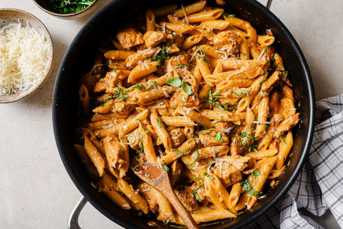 A skillet filled with penne pasta and pieces of chicken in a creamy sauce, garnished with herbs. A wooden spoon rests in the skillet. Nearby are bowls of grated cheese and chopped parsley on a light surface.