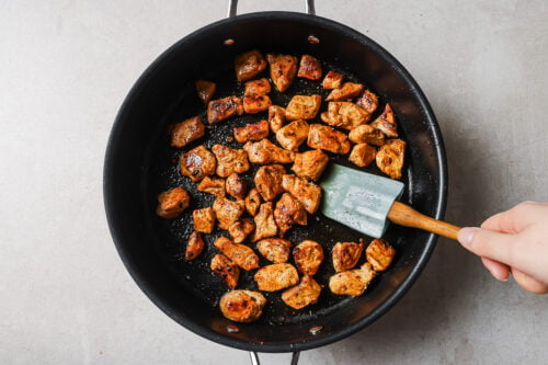 A hand holding a spatula stirs browned, seasoned chicken pieces in a black frying pan on a light-colored countertop.