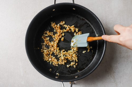 A hand stirs chopped onions in a black skillet with a blue spatula; the onions are sautéing and beginning to turn golden brown.