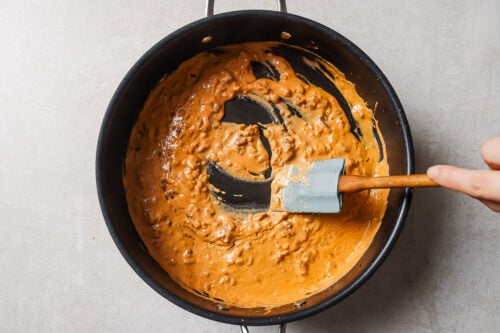 A hand stirs a thick, creamy, orange-brown sauce with a spatula in a black pan on a light countertop.