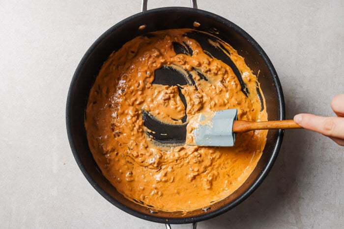A hand stirs a thick, creamy, orange-brown sauce with a spatula in a black pan on a light countertop.