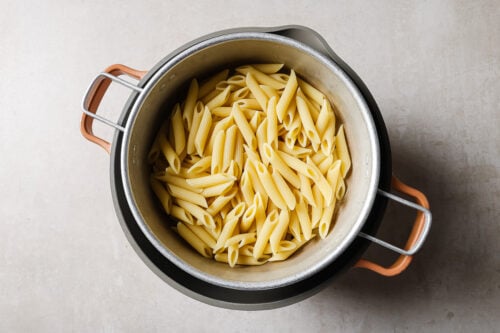 A pot filled with cooked penne pasta, viewed from above, sitting on a light-colored surface.