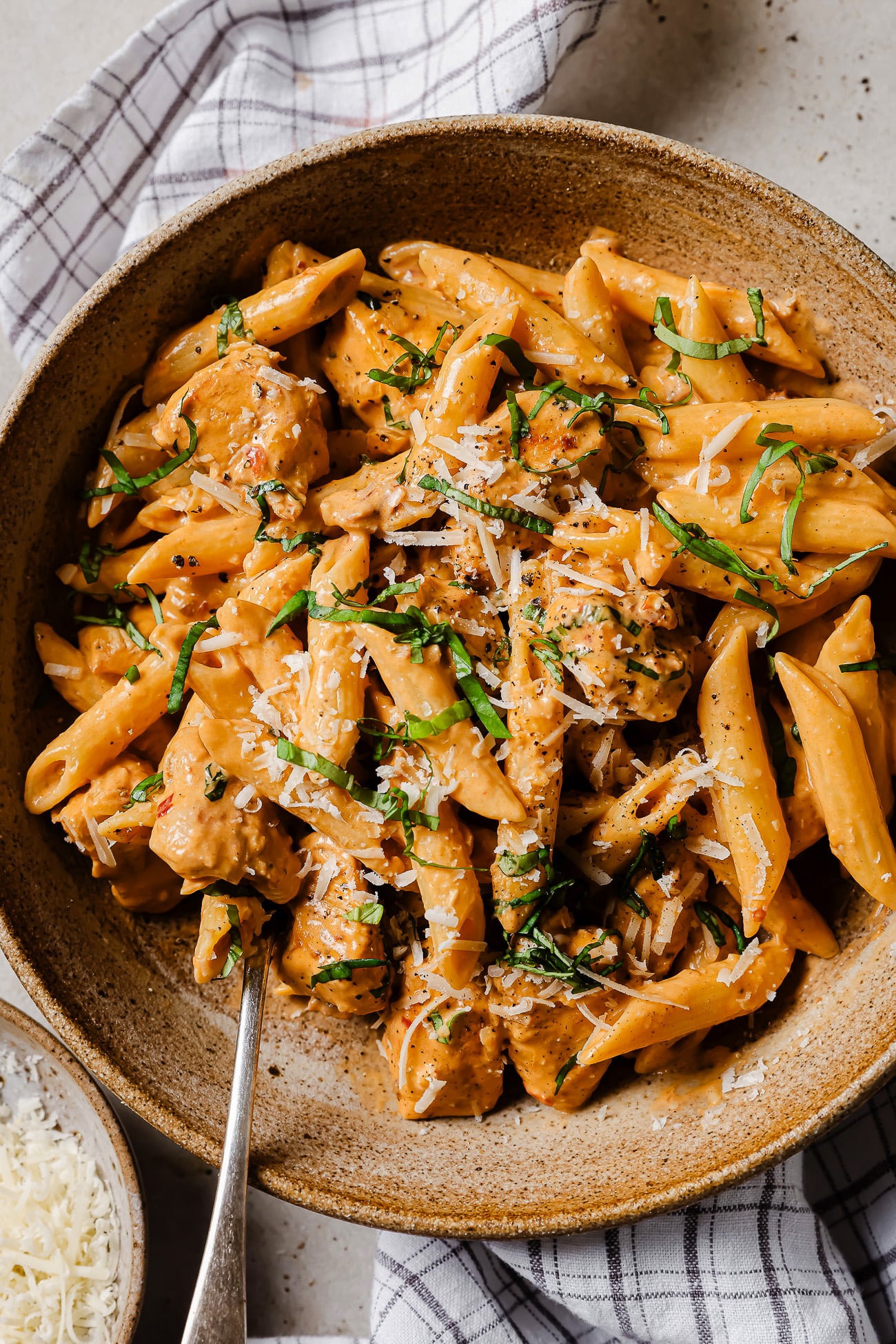 A bowl of creamy penne pasta with pieces of chicken, topped with grated cheese and chopped fresh herbs. A fork is placed in the bowl, and a checked cloth napkin is partially visible underneath.