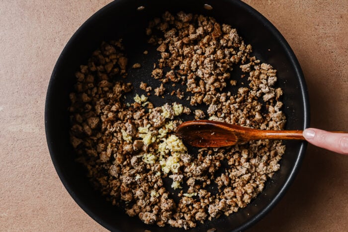 A hand stirs cooked ground meat and minced garlic in a black skillet with a wooden spoon on a brown countertop.