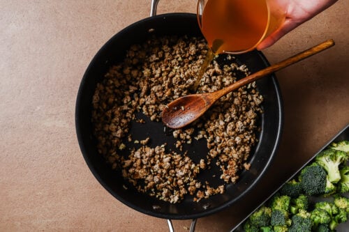 A hand pours broth from a measuring cup into a skillet with browned ground meat, stirred by a wooden spoon. Fresh broccoli is visible on a tray beside the skillet.