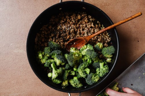 A black pan on a brown surface contains cooked ground meat and fresh broccoli florets being stirred with a wooden spoon. A hand is adding more broccoli to the pan from a metal tray.