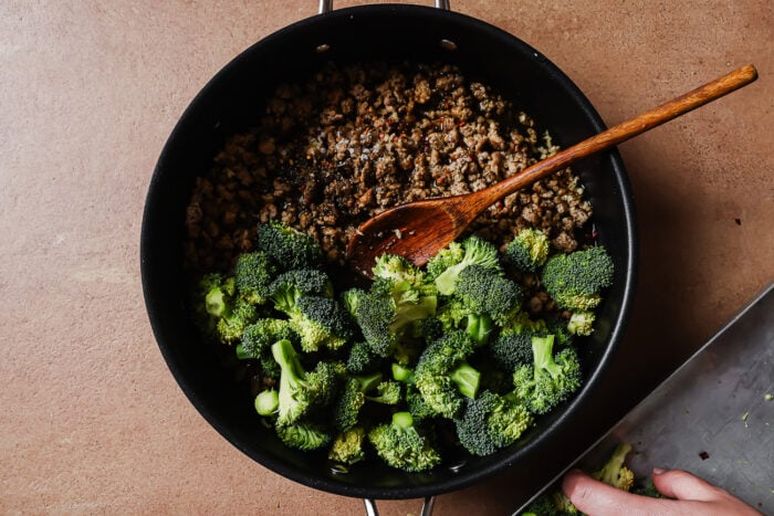 A black pan on a brown surface contains cooked ground meat and fresh broccoli florets being stirred with a wooden spoon. A hand is adding more broccoli to the pan from a metal tray.