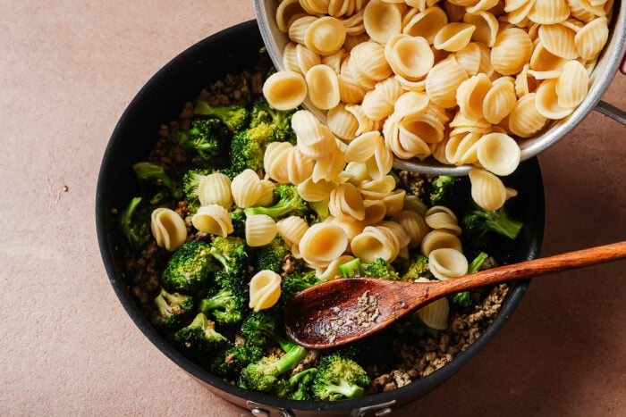 Cooked orecchiette pasta is being poured from a pot into a skillet containing sautéed broccoli and ground meat, with a wooden spoon resting in the mixture.