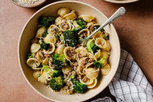 A bowl of orecchiette pasta mixed with cooked ground meat, broccoli florets, and grated cheese, with a fork in the bowl and a checked cloth napkin beside it.