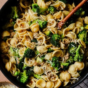 A skillet filled with orecchiette pasta, broccoli florets, crumbled sausage, and grated Parmesan cheese, with a wooden spoon and a bowl of extra cheese nearby on a striped cloth.
