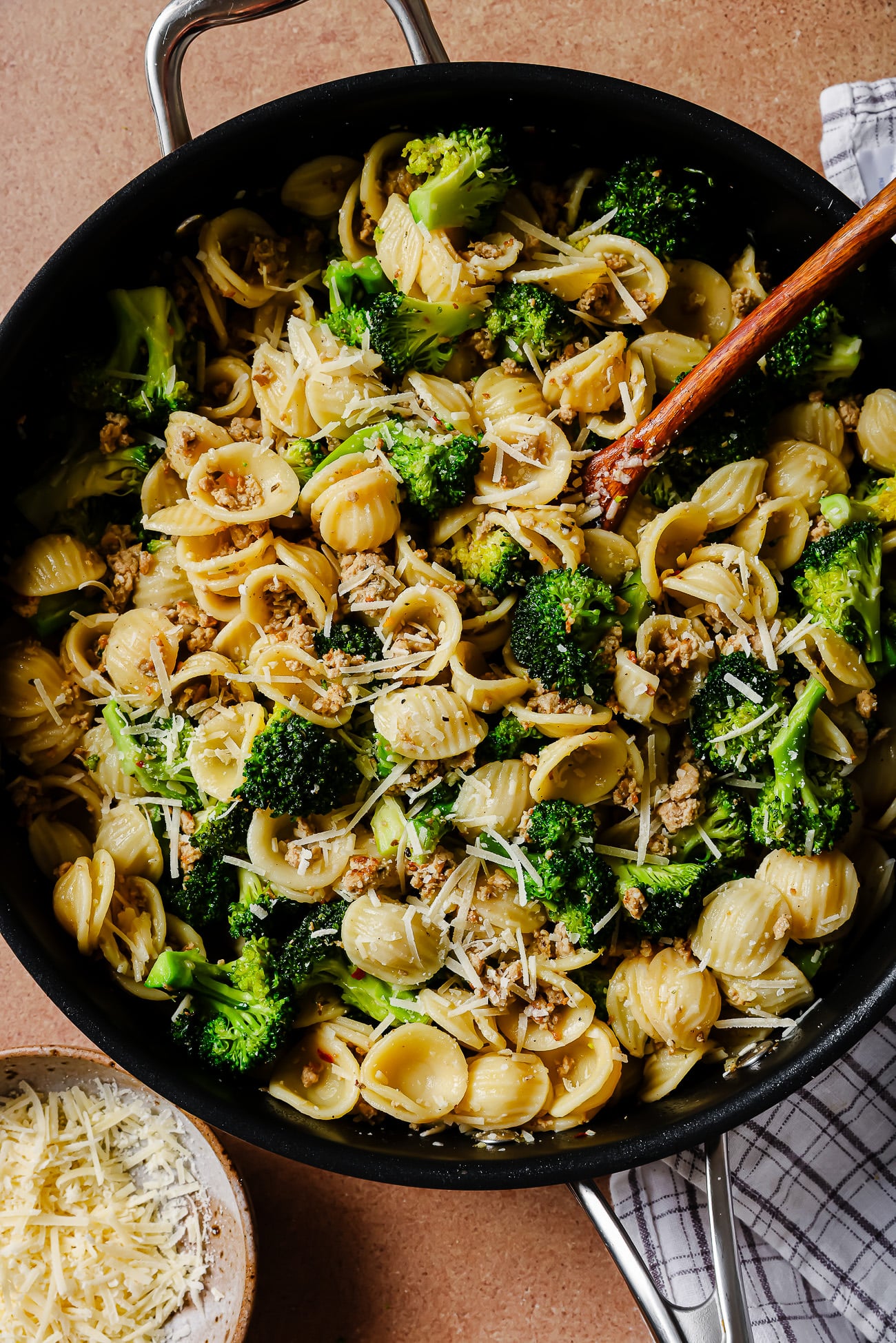 A skillet filled with orecchiette pasta, broccoli florets, crumbled sausage, and grated Parmesan cheese, with a wooden spoon and a bowl of extra cheese nearby on a striped cloth.