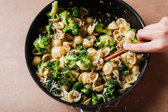 A hand stirs orecchiette pasta with broccoli, mushrooms, grated cheese, and ground meat in a black skillet on a light brown surface.